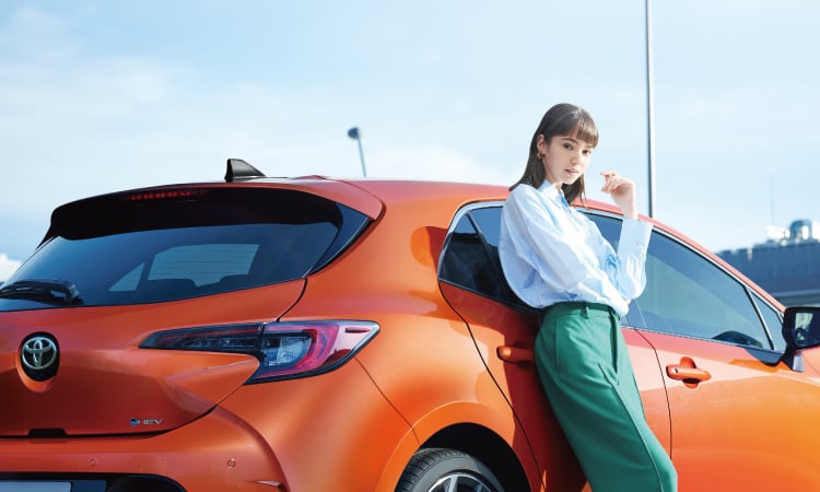 カローラ スポーツ G“Z”(ハイブリッド)。ボディカラーはオレンジマイカメタリック。車の横に立つ女性の様子。