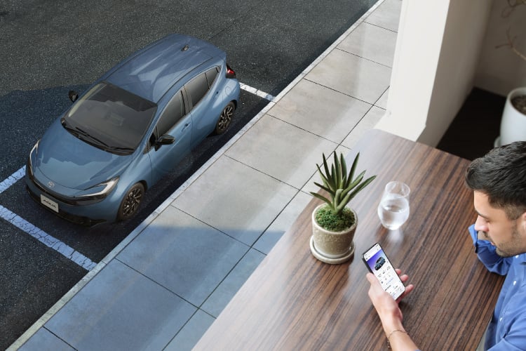カフェの席からスマートフォンを操作する男性と窓の外の駐車場に停まるアクア Z（2WD・ボディカラーはグレイッシュブルー）